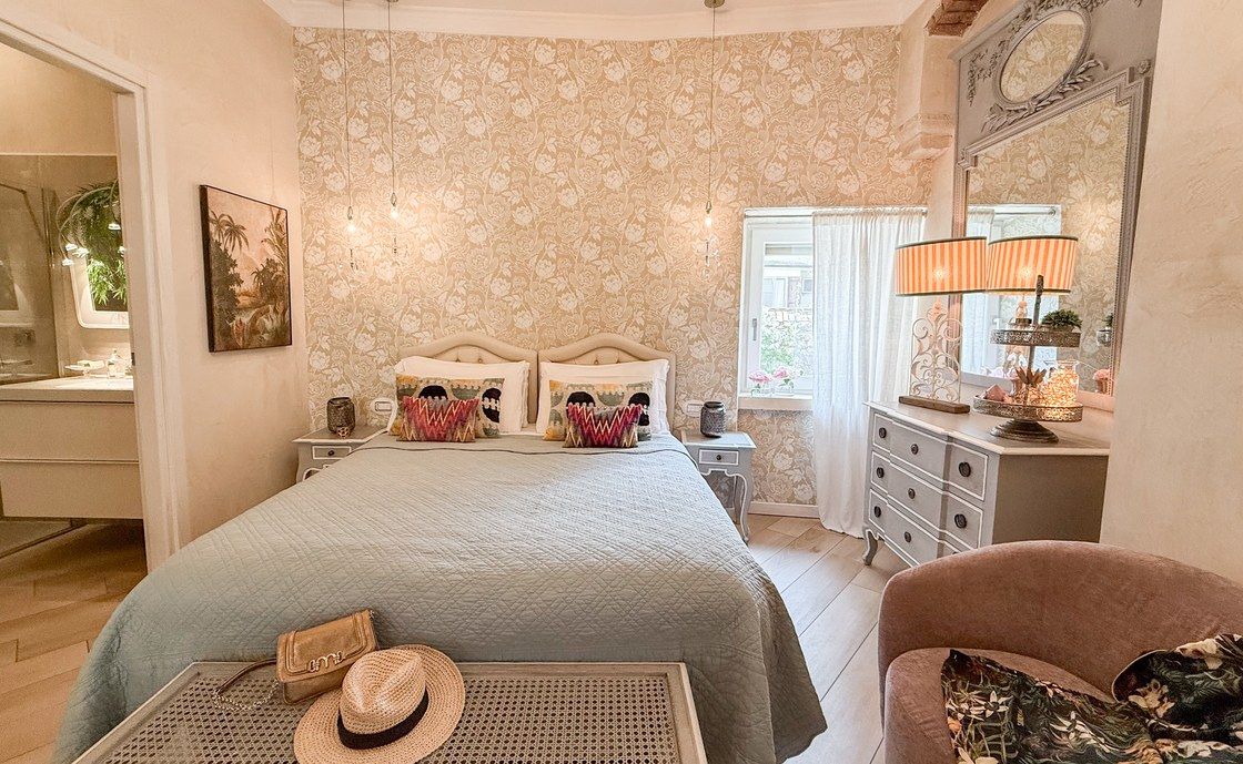 Bedroom at Santo Stefano with floral wallpaper, a sage green bedspread and a straw hat on the cane bench
